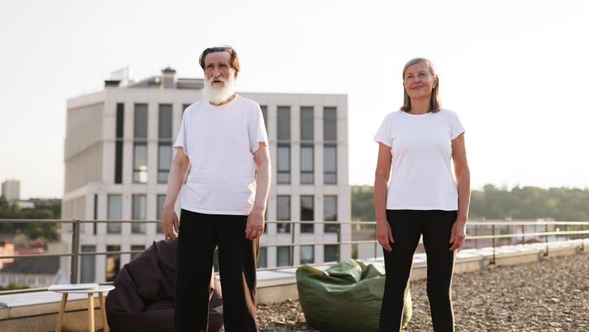 Older man and woman performing workout together on rooftop terrace, focusing on squats and physical health. Engaging in active lifestyle for improved fitness and well-being.