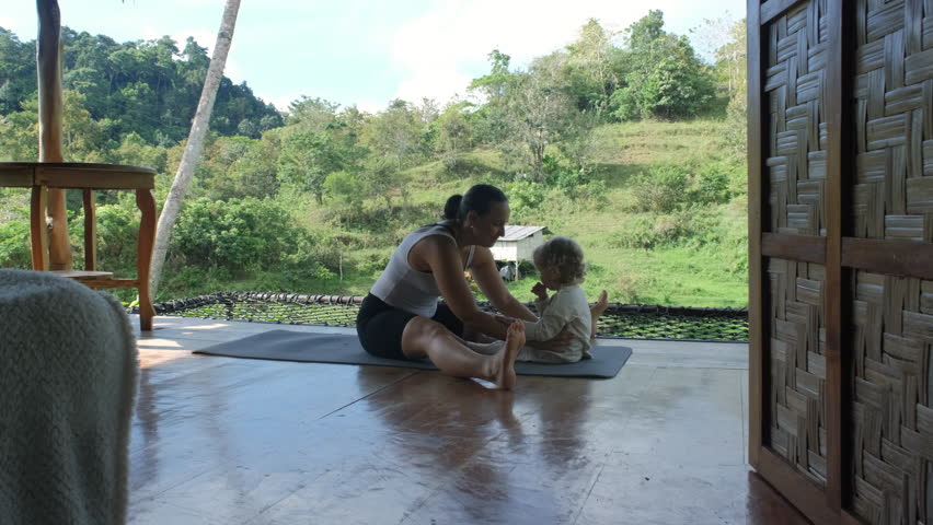Young woman engages with her daughter while exercising on a terrace of hut amidst amidst lush tropical greenery and rice plantations, slow motion
