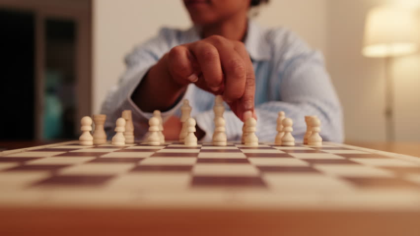 A dark-skinned person sets up white chess pieces on a chessboard in a medium close-up shot. They carefully position each piece, ready to start a game.