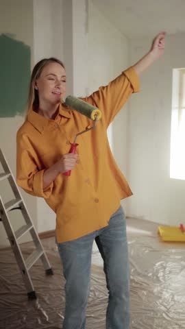 Woman engaged in home renovation, using a roller brush to paint walls. A ladder and paint supplies are visible, indicating an ongoing diy project in a bright, airy room
