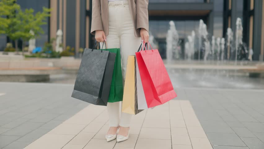 Outdoor city shopping moment with a fashionable woman holding several brightly colored shopping bags, standing near a large public water fountain.