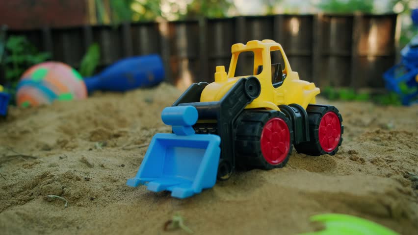 A bright yellow bulldozer toy digs in the sand at a playground filled with colorful plastic toys The scene captures fun outdoor playtime under clear blue skies and sunlight