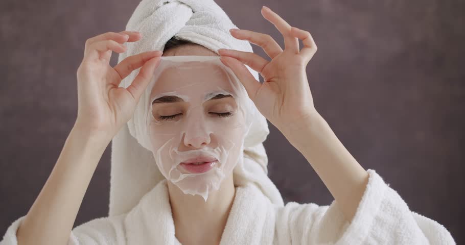 Beautiful woman in a bathrobe and a towel on head applying cosmetic sheet mask on face at home. Woman wearing paper sheet facial mask ready for moisturizing anti wrinkle day or night cream at home.