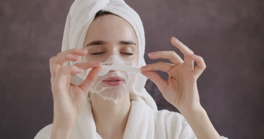Beautiful woman in a bathrobe and a towel on head applying cosmetic sheet mask on face at home. Woman wearing paper sheet facial mask ready for moisturizing anti wrinkle day or night cream at home.