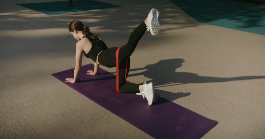 Athletic brunette female in sportswear exercising, stay in plank with resistance band and raise leg up. Sporty woman have workout on exercise mat outdoors. Woman doing yoga plank pose on yoga mat.