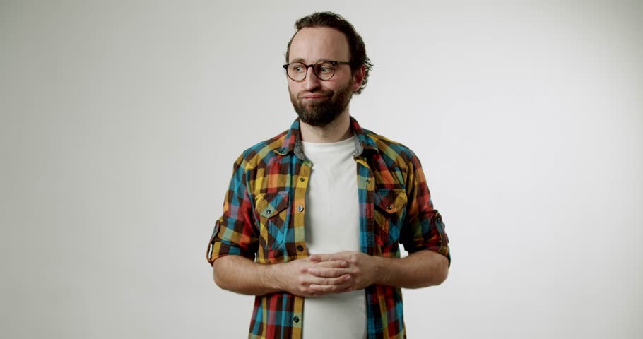 A bearded man in glasses and a plaid shirt stands against a white background, looking displeased and holding his hands clasped in front of his stomach. He appears to be thinking or waiting.