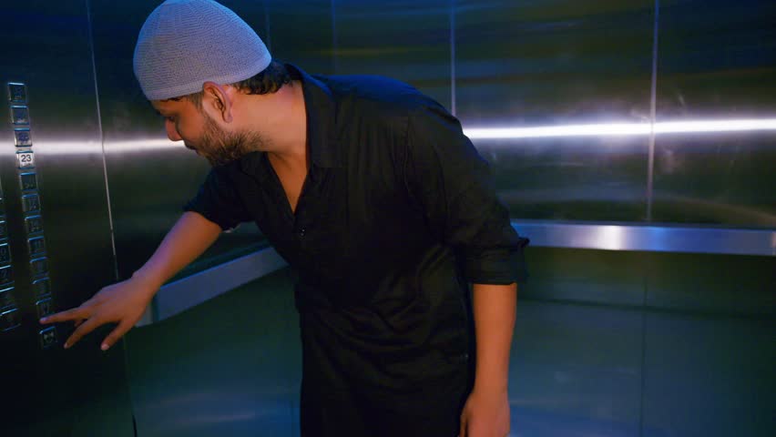 Muslim man taps lift panel in silent confusion, Puzzled guy tries elevator switch once again, Uncertain traveler hits floor key nervously, Male with kufi represses button quickly, Person check numbers