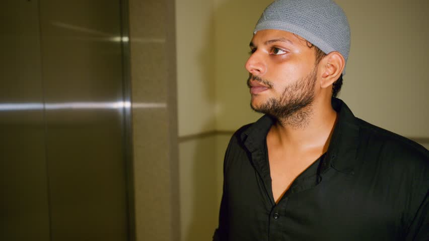 Puzzled guy tries elevator switch once again, Muslim man taps lift panel in silent confusion, Uncertain traveler hits floor key nervously, Male with kufi represses button quickly, Person check numbers