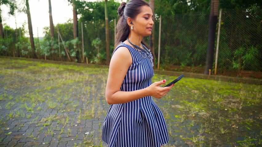 Fashion girl in mini attire enjoys mobile tune, Young lady strolls by garden syncing with melody, Chic walks slowly humming along digital sound, Stylish female grooves gently listening via earphone