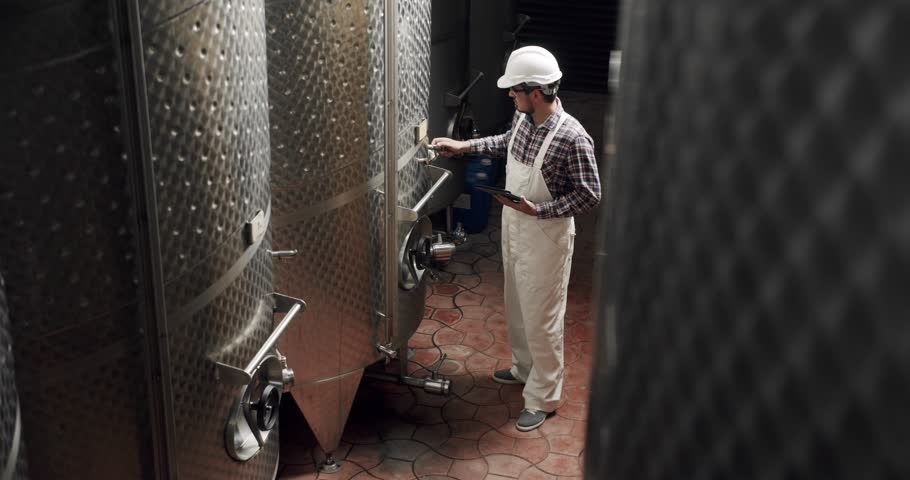 Male wine maker checking quality of beverage distillation process indoors and write down the dates in the diary. Alcohol production concept.Controls the quality of production equipment in wine factory