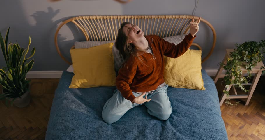 Energetic and cheerful girl in casual clothes sits on the bed and listens to energetic music plays as if on a guitar and imagines it at home in a modern apartment