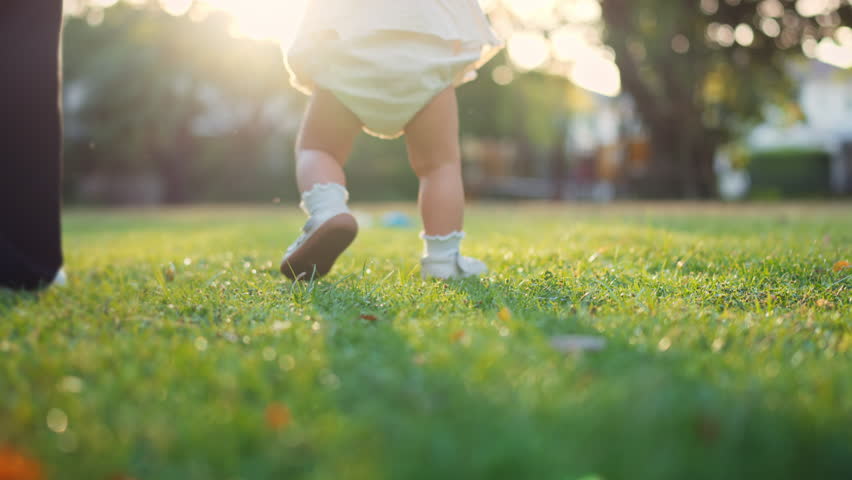 Close-up of baby wearing shoes taking first steps on green grass in park with soft morning sunlight – toddler walking with mother, childhood milestone and outdoor lifestyle concept