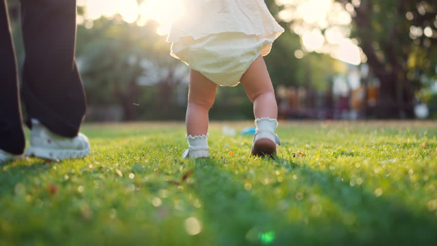 Close-up of baby wearing shoes taking first steps on green grass in park with soft morning sunlight – toddler walking with mother, childhood milestone and outdoor lifestyle concept