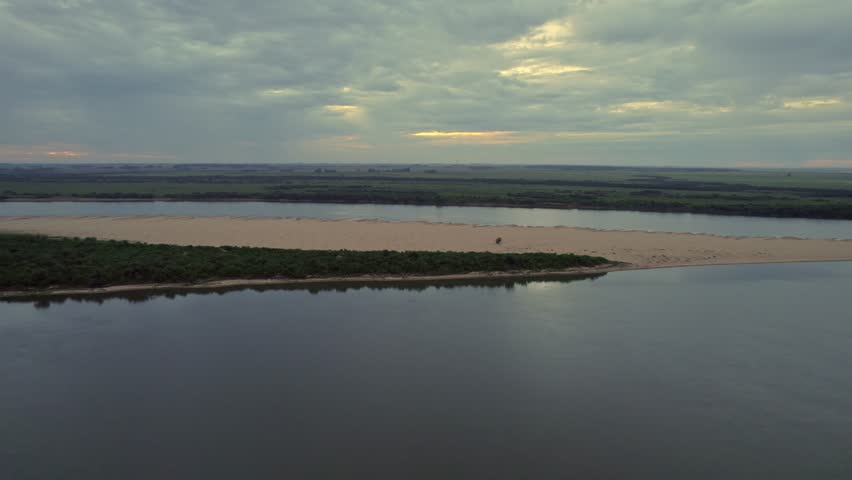 An aerial landscape view of a sandbank with greenery surrounded by water, and field in the background under cloudy sky at sunset