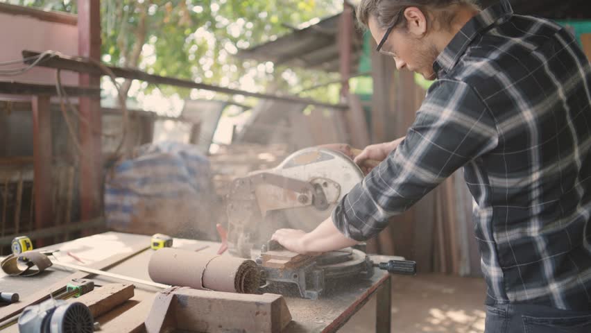 Male carpenter wearing protective eyewear using electric circular saw cutting wood board at workshop studio