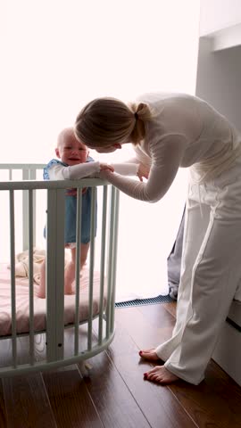 Crying baby standing in crib while mother gently comforts her. Real parenting moment toddler tantrum, emotions, love and care at home. Authentic motherhood, daily life, baby behavior indoor