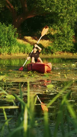 Male tourist is kayaking in red canoe along calm river at sunset. Young man floats in boat on lake in summer. Active tourist is rafting in kayak. Vertical video