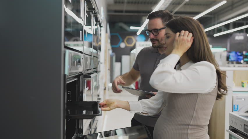 Friendly salesman holding a digital tablet is showing different modern ovens to a pregnant customer in a home appliances store, they are both smiling and looking at the oven