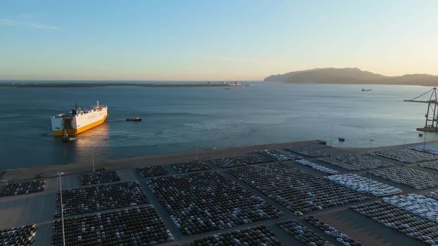 Ro-Ro Cargo Ship with Tugboats Arriving and Rows of New Cars on Large Full Parking Lot at Sunset in Port. Automotive Industry. Setubal, Portugal. Aerial View. Moving Forward