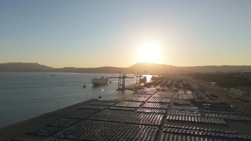 RORO Cargo Ship and New Cars on Large Full Parking Lot at Sunset in Port Ready for Distribution. Car Storage, Automotive Industry. Ocean and Mountains. Setubal, Portugal. Aerial View. Moving Forward