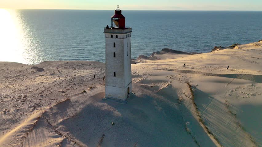 Explore the stunning landscape of Rubjerg Knude Fyr Lighthouse in Jutland, Denmark, as warm sunset light bathes the iconic lighthouse.