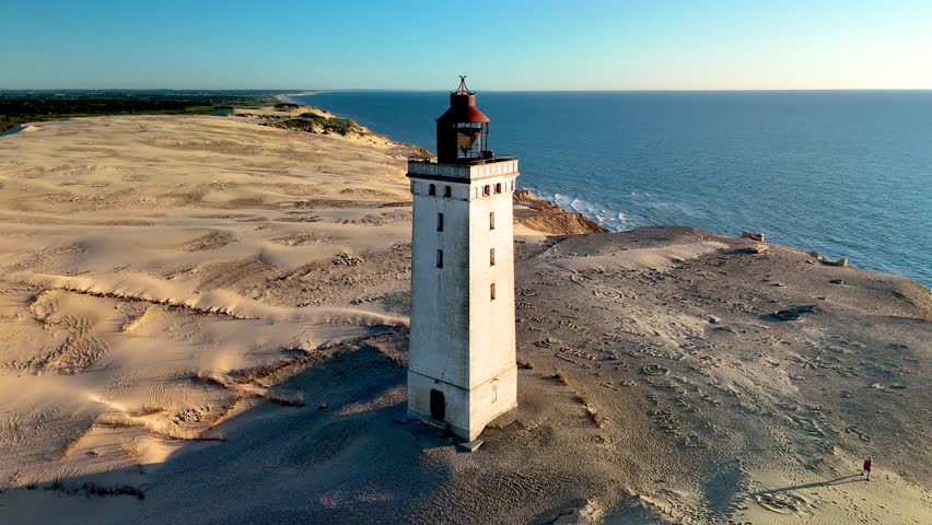 Discover the breathtaking beauty of Rubjerg Knude Fyr Lighthouse in Jutland, Denmark, featuring golden sands, gentle waves, and a majestic lighthouse against a vibrant sunset sky, 