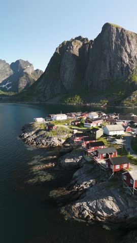 A picturesque coastal village in Hamnoy Lofoten Norway features traditional red wooden houses along the rocky shoreline, surrounded by towering mountains.
