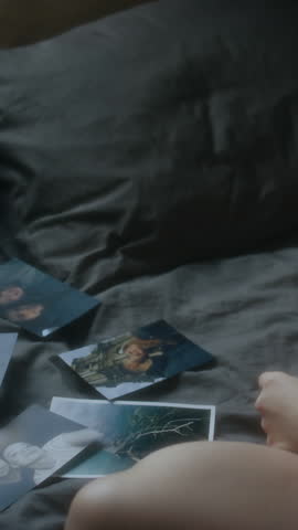 Vertical shot of unrecognizable woman sitting in bed and looking through pictures with boyfriend, tearing one photo out of harsh pain from breakup