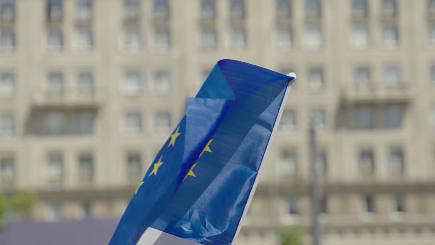 European Union flag attached to a pole, waving in the wind in an urban setting, possibly during a peaceful protest. Symbol of unity, democracy, and European identity.