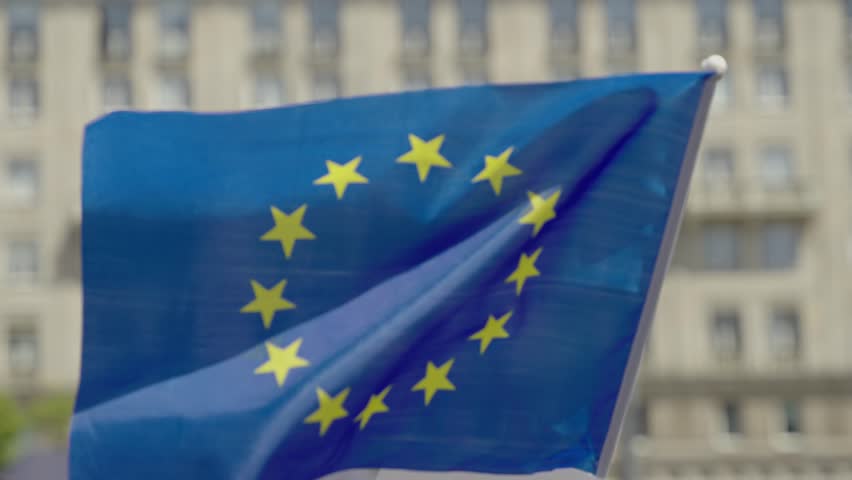 European Union flag attached to a pole, waving in the wind in an urban setting, possibly during a peaceful protest. Symbol of unity, democracy, and European identity.