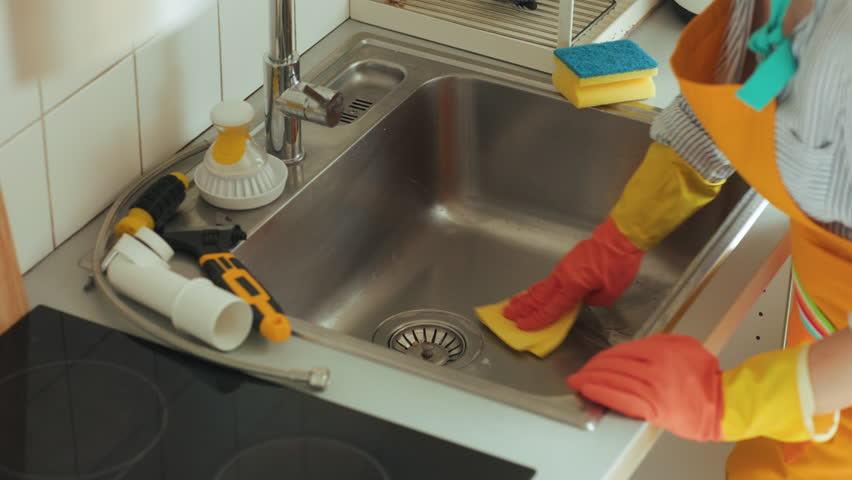 A woman wearing an orange apron and colorful gloves cleans a stainless steel sink with a yellow sponge after plumbing repairs. Tools and cleaning solution are visible.