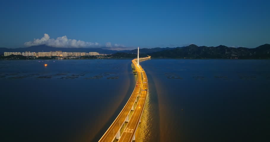 Aerial shot of an illuminated modern bridge spanning across the bay with city lights and mountains at night in Shenzhen, China.