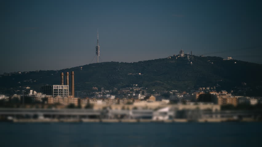 Nighttime establishing shot of Barcelona port features a blurred sailboat, city landmarks, distant hills, and telecom antennas.
