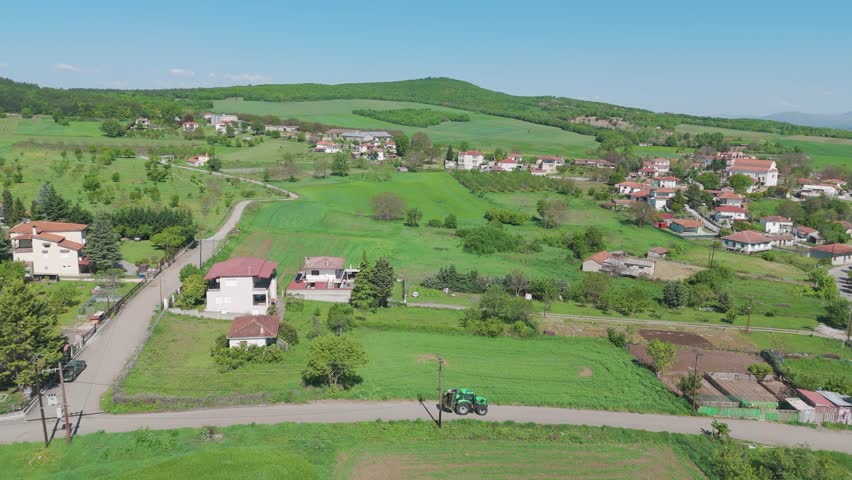 Tractor roaming through village surrounded by green fields, scattered houses, and rolling hills under clear blue sky, drone shot