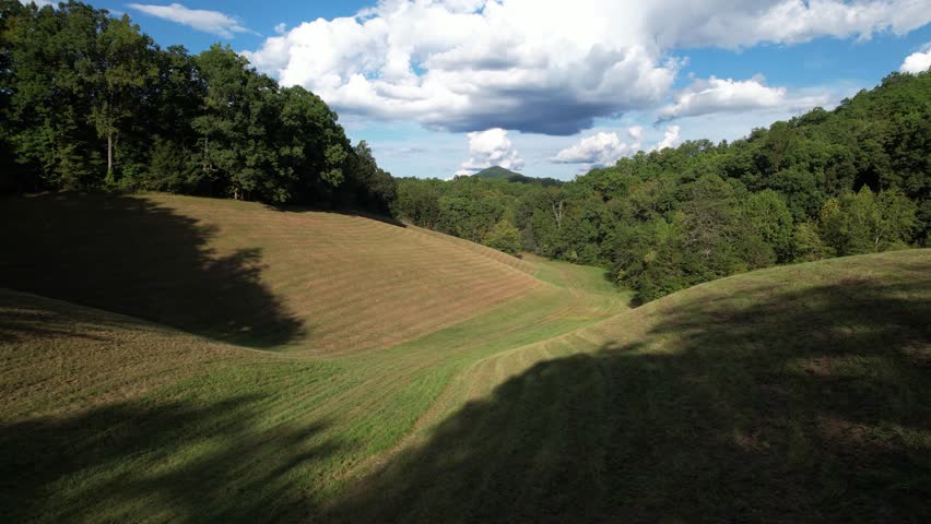 Smooth zoom-in over an open grassy field bordered by dense forested hills under a partly cloudy sky in a rural landscape.
