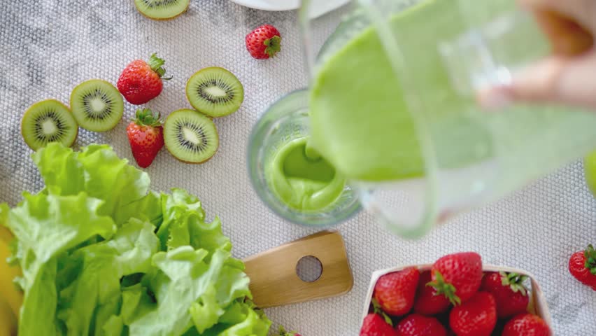 Top view of a green smoothie being poured into a glass, surrounded by fresh kiwi, lettuce, and strawberries on a white textured table. The vibrant colors and healthy ingredients, 4k 60fps slow motion.