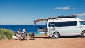 Mother and daughter relaxing in foldable chairs beside their camper van, admiring a stunning ocean view under a clear blue sky. - Powered by Shutterstock - Get 15% off with code: PIKWIZARD15