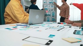 A diverse team collaborates around a table filled with design materials, laptops, and colorful sketches, focusing on creating a mobile app in a bright office setting. SACTR - Powered by Shutterstock - Get 15% off with code: PIKWIZARD15