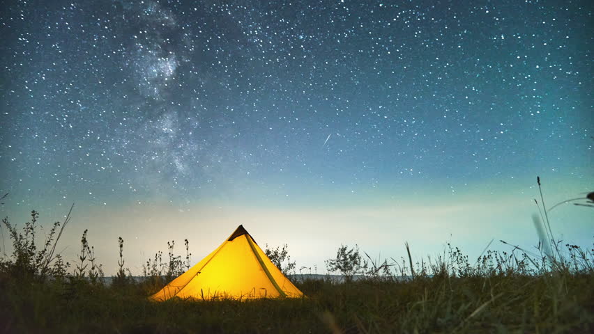Time lapse of grassy hill with illuminated camp tent under moving night sky with stars. Beautiful view of night starry sky over tourist tent in mountains. Hiking, traveling and night camping concept.