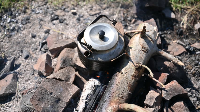 Close-up of metal kettle heating over campfire. Flames and smoke curl around well-used kettle, which rests on logs. Blur of green grass, emphasizing rustic outdoor setting on background.