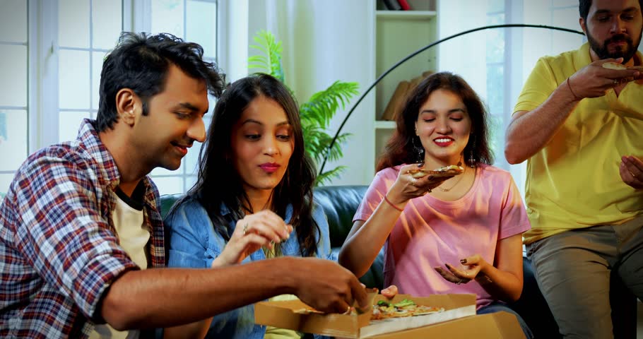 Friends having pizza at home, Indian Asian young close friends sitting together on sofa in cozy modern house, smiling and enjoying cheesy slices while showing pizza box or slice to camera with joy