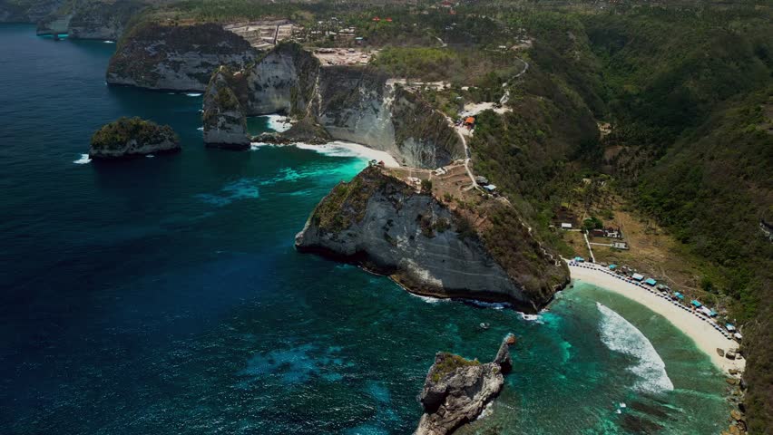 Aerial drone footage of Diamond Beach in Bali shows towering white cliffs and clear blue ocean as sunlight reflects off the water and visitors explore the shore.