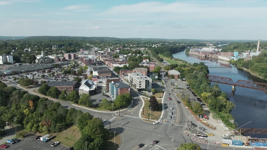 Aerial view of downtown Waterville, Maine showing city streets, modern buildings, the Kennebec River, and a historic railway bridge on a clear summer day.