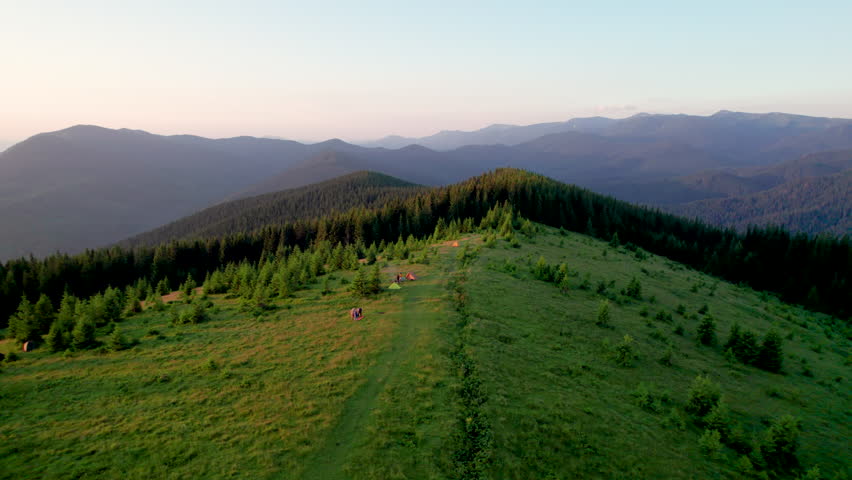 Aerial view of morning camping. Campsite at sunset with tourist tents among evergreen trees. Campers sit nearby, facing vibrant sunset over rolling hills. Serene and picturesque scene.