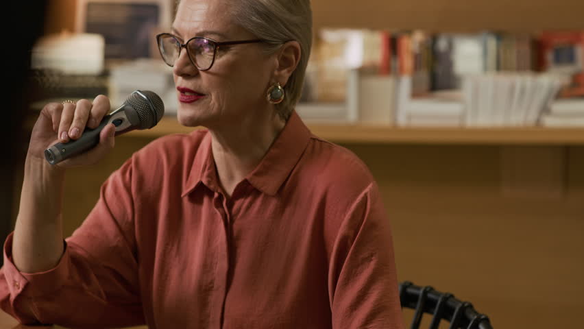 Confident mature Caucasian female author speaking into microphone during intimate bookstore event, sharing insights and engaging attendees in literary discussion