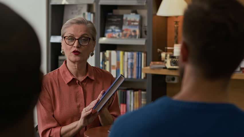 Confident female author holding up her latest book while speaking to small group of readers during bookstore event, answering their questions in warm and friendly manner