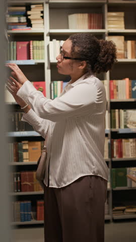 Vertical medium shot of young Biracial woman standing in bookstore aisle, thoughtfully flipping through book among tall shelves in a calm, literary atmosphere