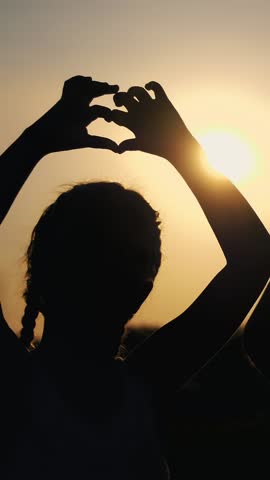 Silhouettes, figures of children, boy and girl show the hearts with the fingers, hands, against the background of the sun, at sunset in summer. Happy family. Slow motion