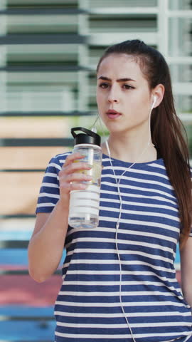 Vertical Video of Sports woman is drinking water from shaker on the background of sports equipment. Sports and healthy lifestyle. Attractive woman at an outdoor workout in a park on the sports field.