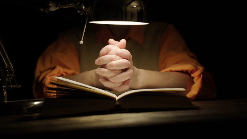 Woman praying with crossed hands over scripture book under table lamp in dark room close up